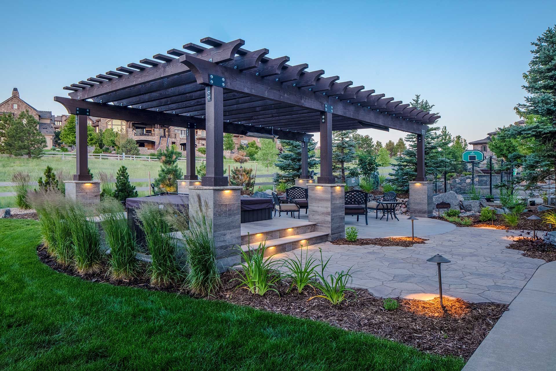 A wooden pergola stands over a stone patio with outdoor furniture, landscape lighting, and a lawn at dusk.