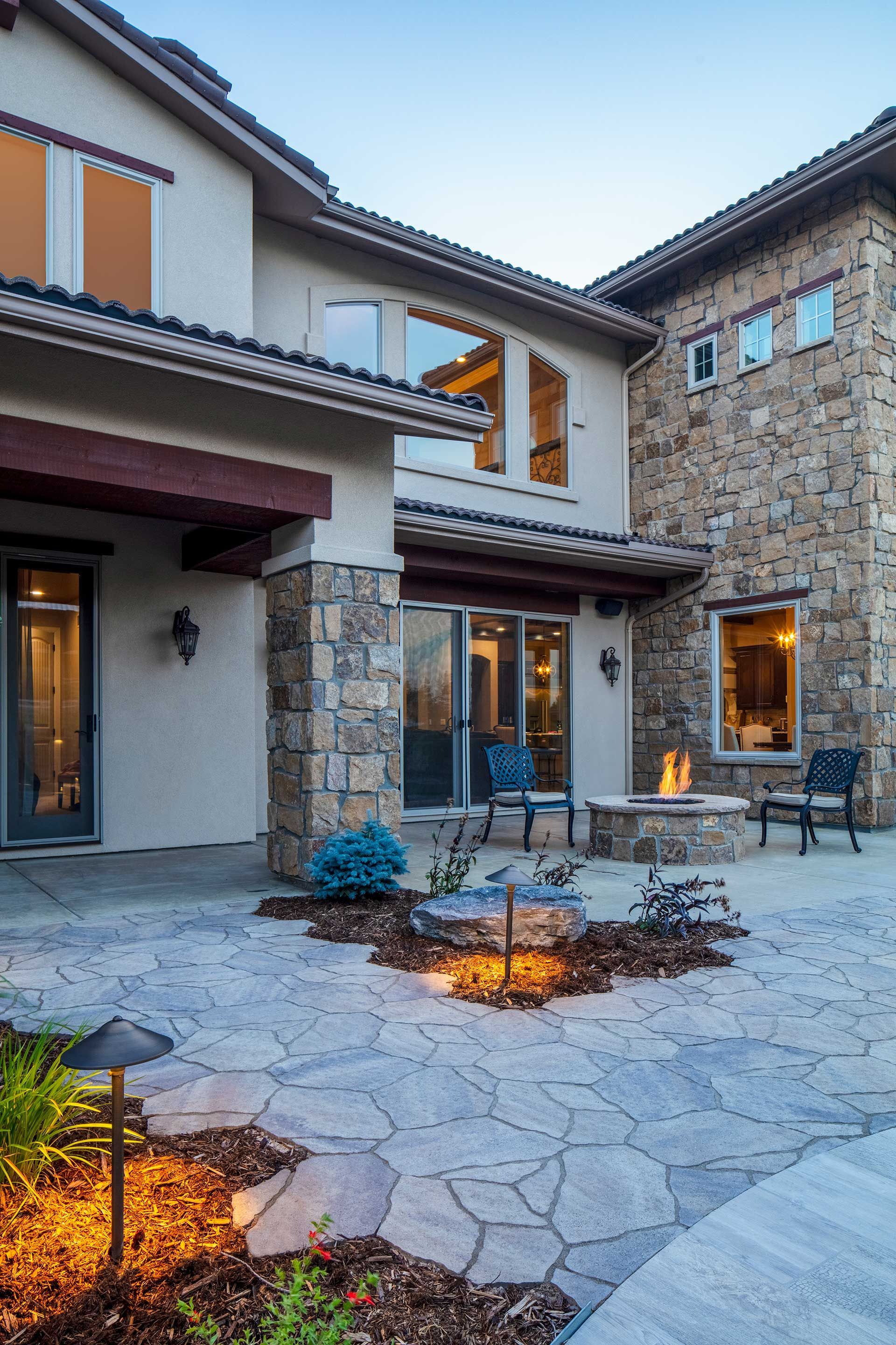 A patio with stone pavers, a fire pit, outdoor chairs, and garden lights at the side of a two-story home with stone siding.