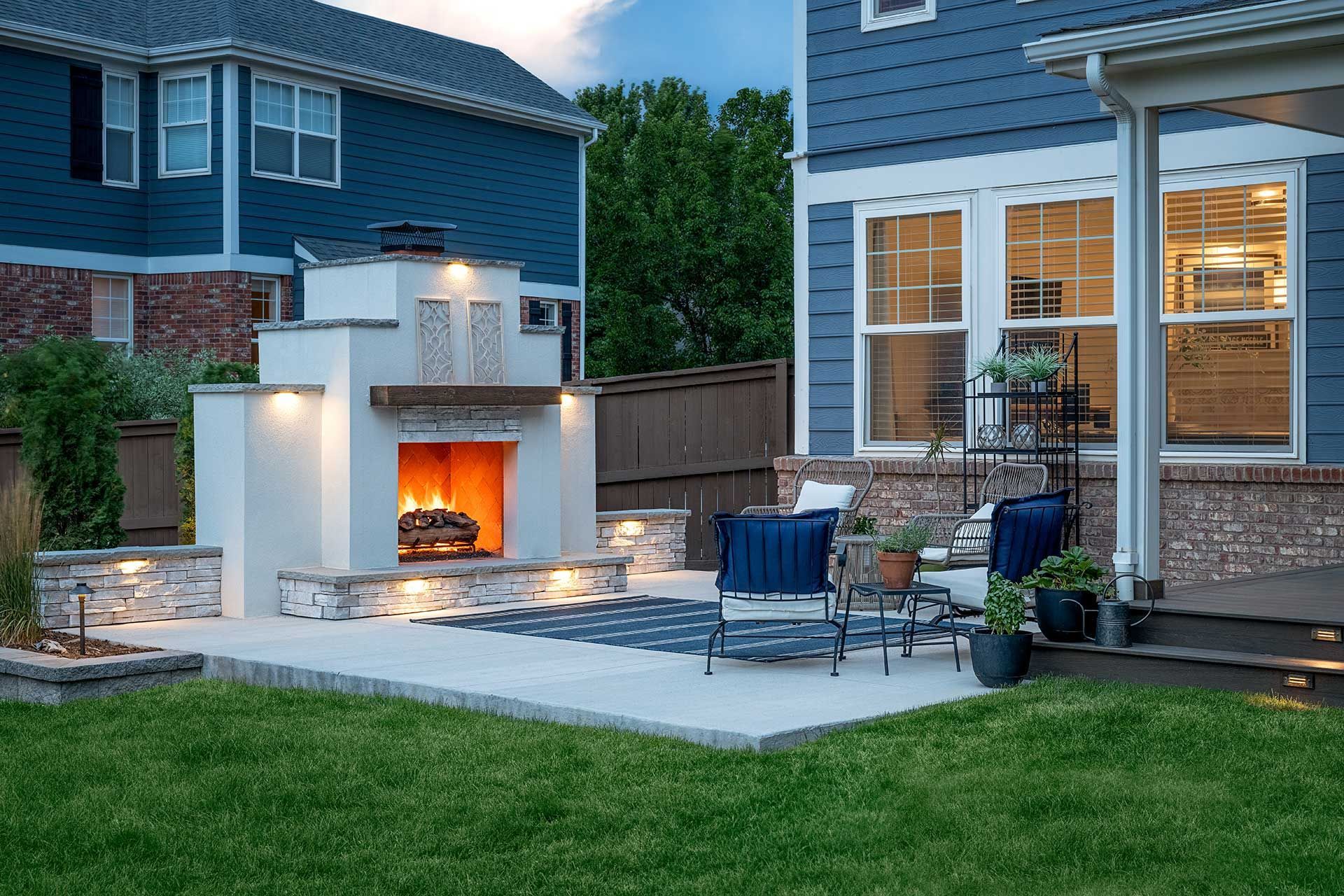 Backyard patio with a stone fireplace, outdoor seating, and a covered porch attached to a blue two-story house at twilight.