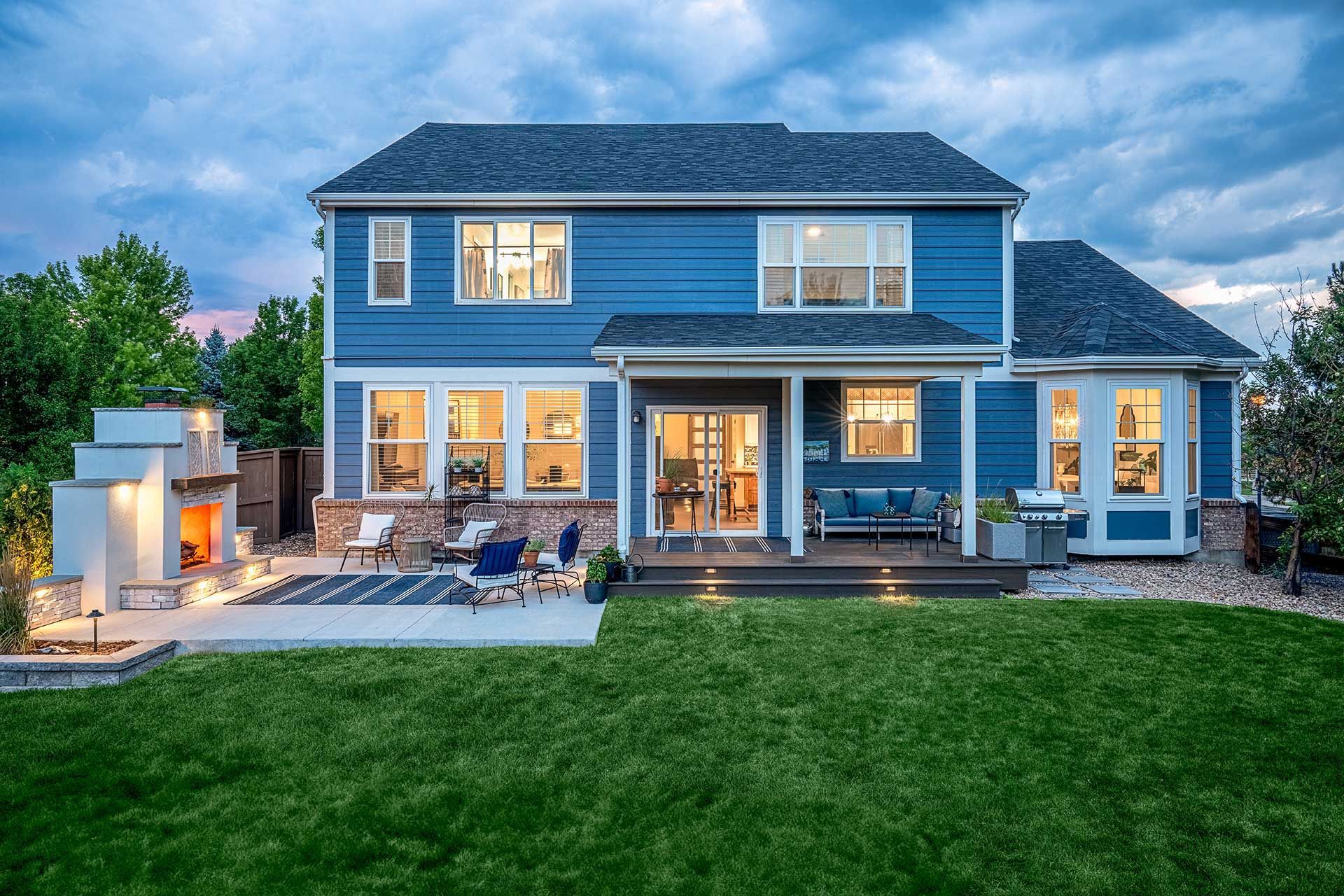 A blue two-story house with a backyard, patio, stone fireplace, and green lawn at dusk.