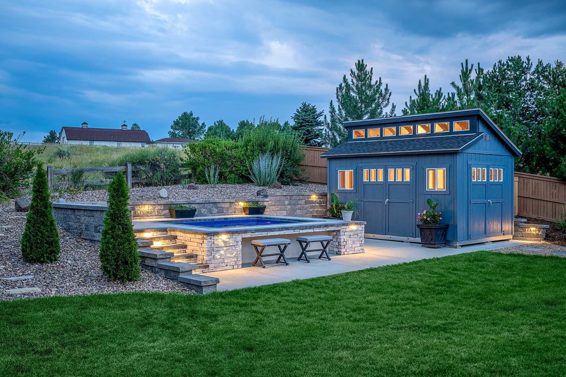 An illuminated stone hot tub with stools sits next to a blue backyard shed on a lawn at twilight.