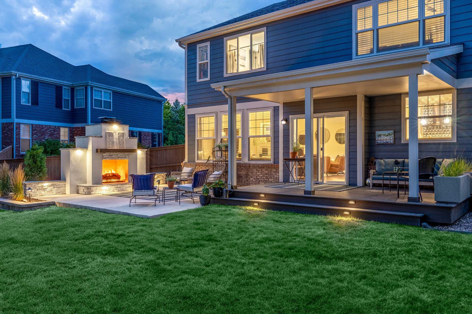 A blue house with a covered patio, outdoor fireplace, and green lawn at dusk.