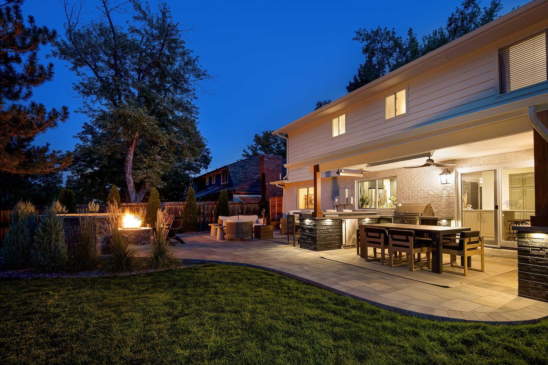 A backyard patio at dusk featuring a lit fire pit, an outdoor dining area, and the brightly illuminated house exterior.