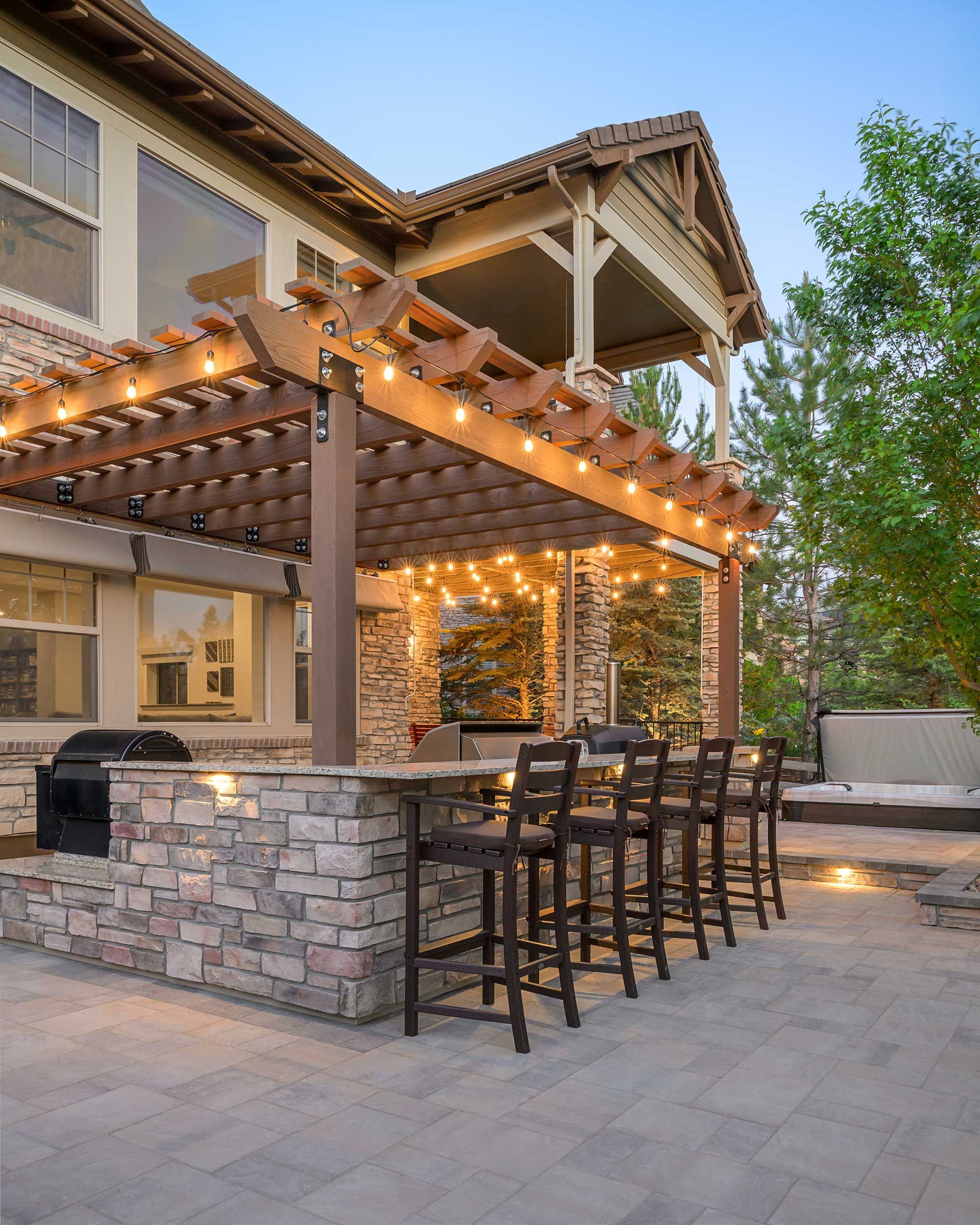 Stone outdoor kitchen and bar with wooden pergola, string lights, and high chairs on a paver patio next to a house.