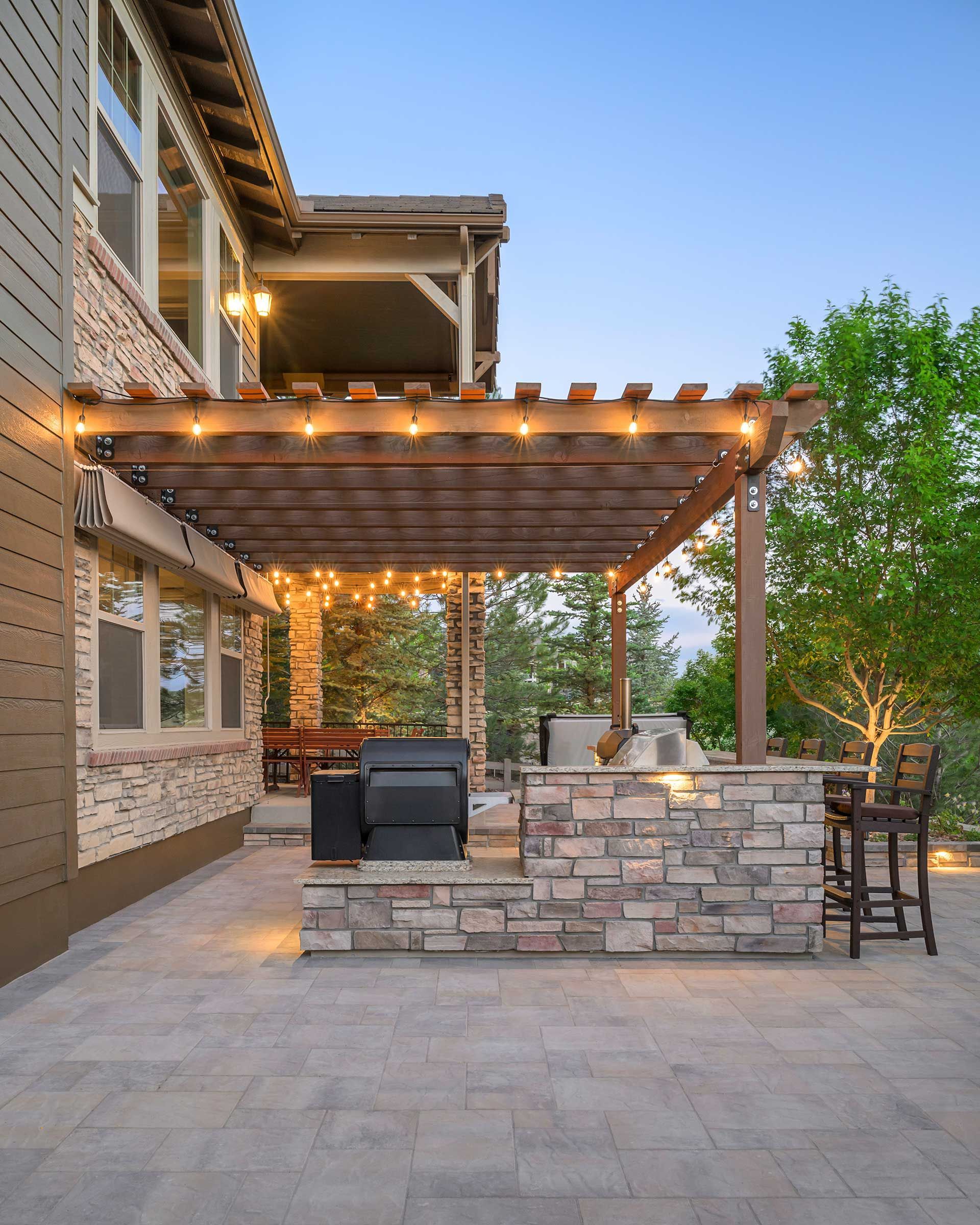 A stone patio featuring a wooden pergola with string lights, a built-in outdoor kitchen, and bar seating at dusk.