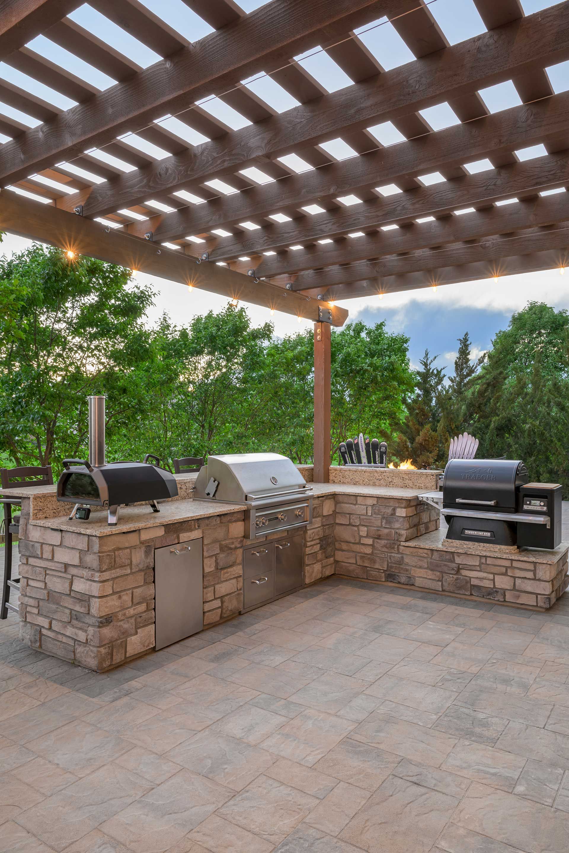 Outdoor kitchen featuring a stone-clad grill station and pizza oven under a wooden pergola on a paved patio.