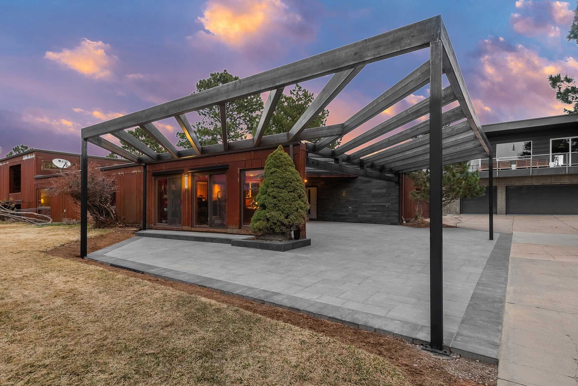 A patio with a large, angled wooden pergola, featuring modern pavers and glass doors against a sunset sky.