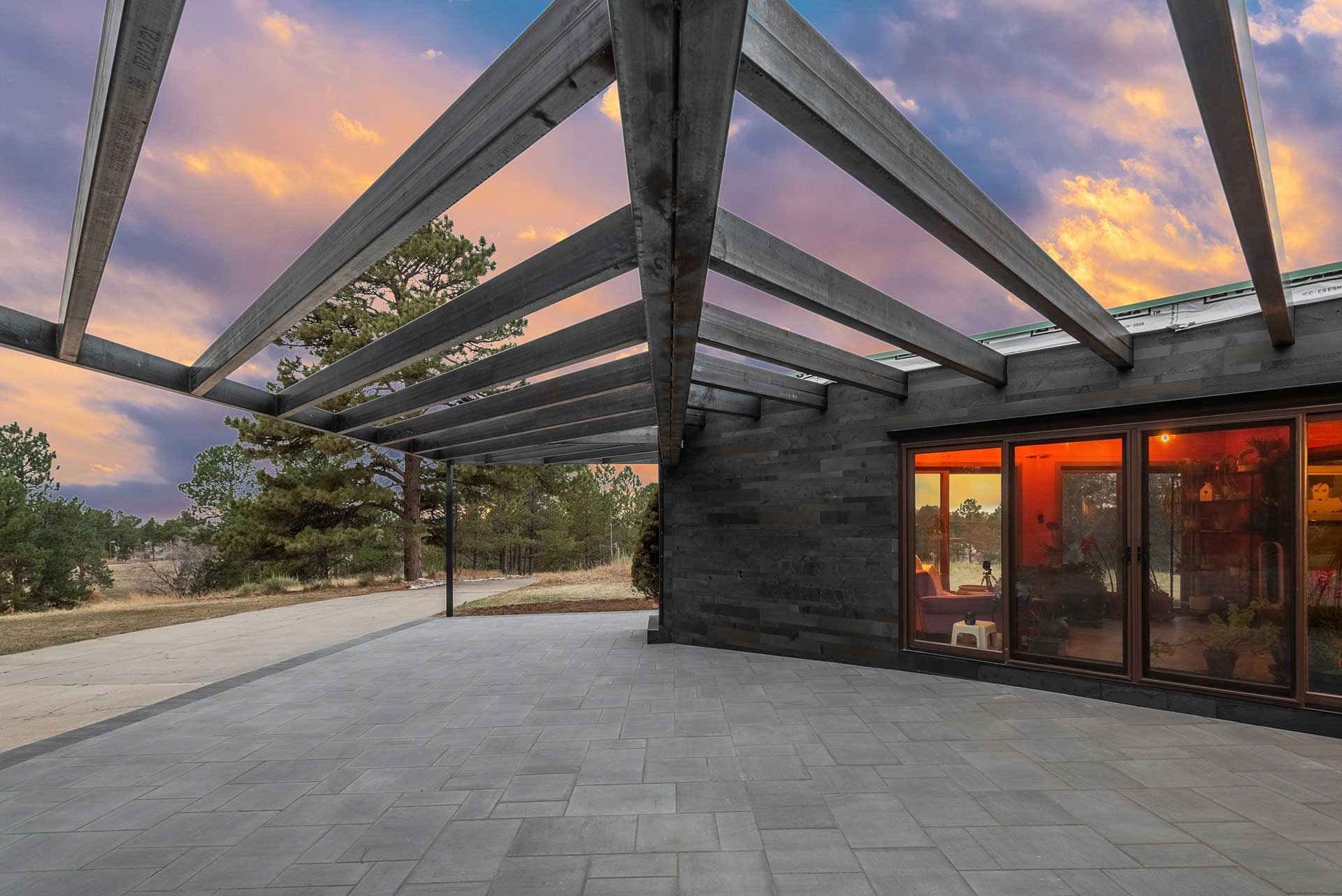 A modern patio with gray pavers and an open wooden pergola under a vibrant sunset sky.