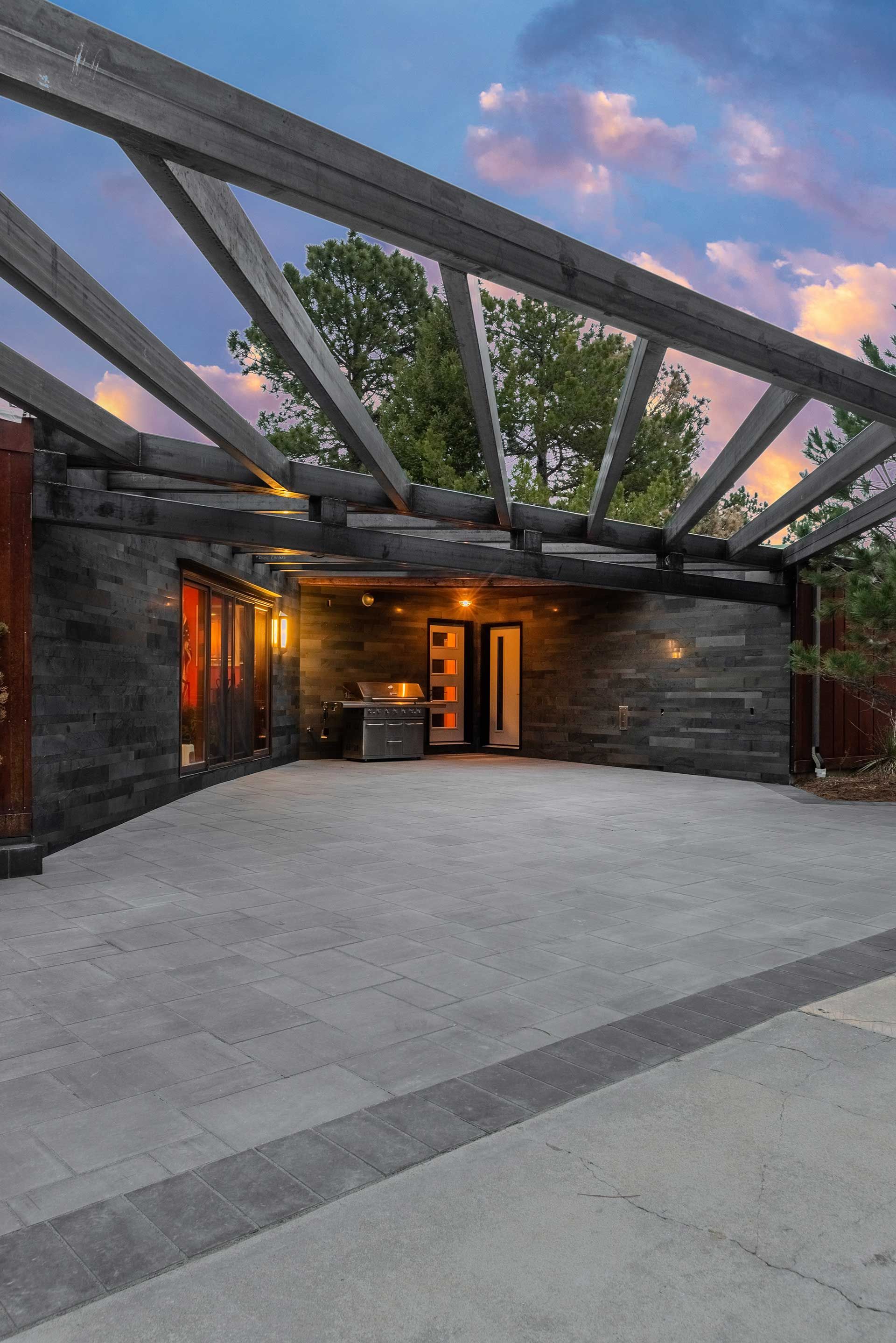 A modern home entrance with dark stone walls, a gray paver patio, and a dramatic arched wooden pergola at sunset.