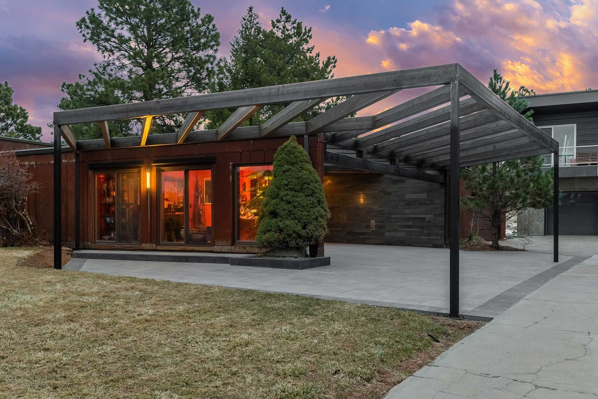 A modern house exterior at sunset, featuring a wooden pergola over a concrete patio and a lit sliding glass door.