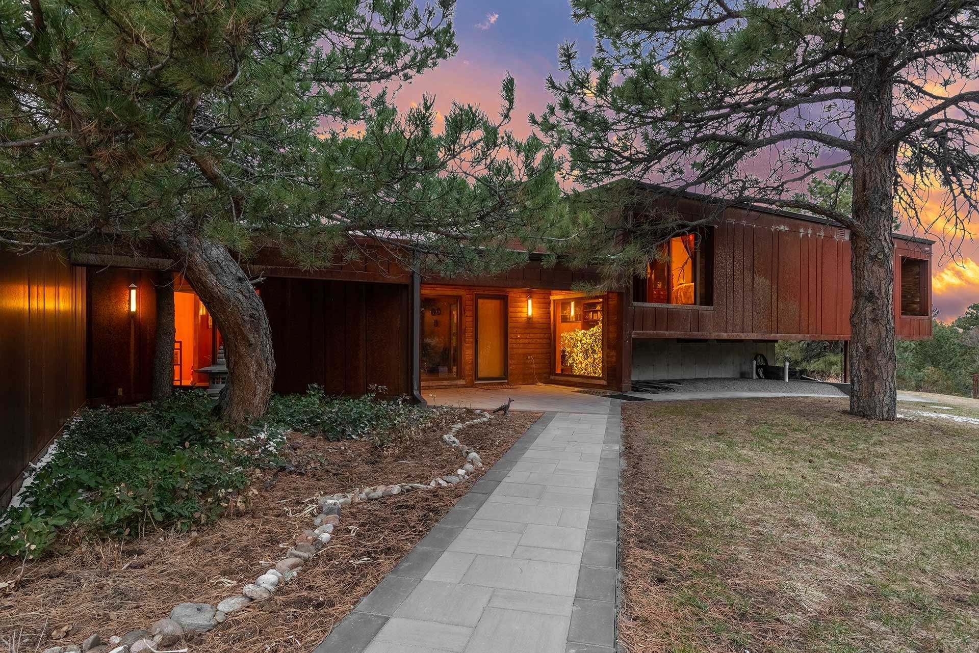 A modern home with wood siding and a stone walkway, surrounded by pine trees under a colorful sunset sky.