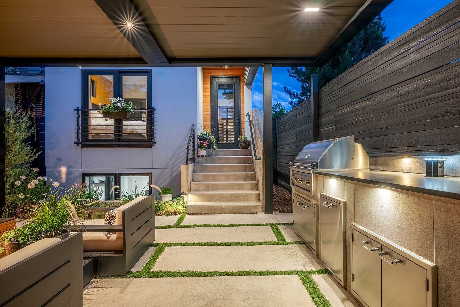 An outdoor patio at dusk with a stainless steel grill station, a stepped entryway, and modern landscaping.