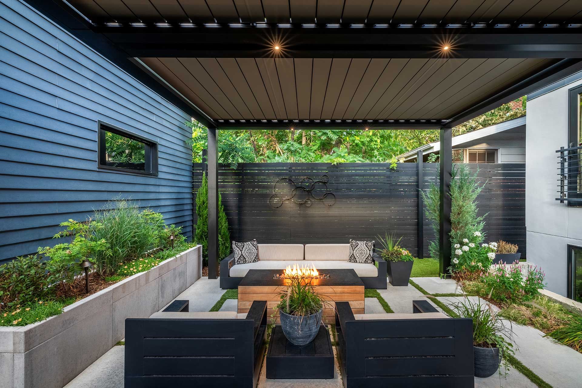 A modern outdoor lounge with a pergola, fire pit, couch, and chairs, set on a stone patio with planters and greenery.