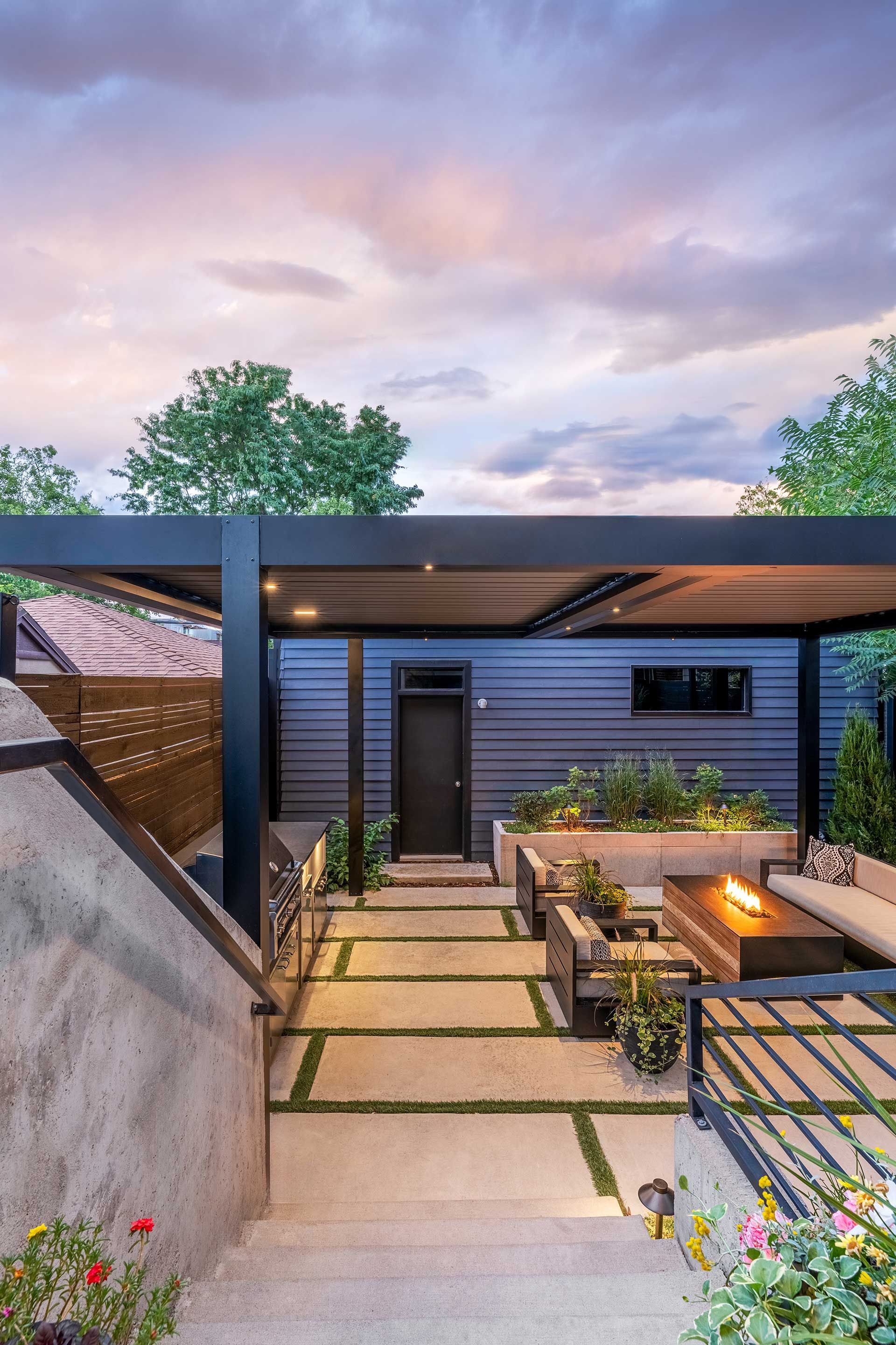 A modern outdoor patio at dusk with a large black pergola, stone pavers, comfortable seating, and a fire pit.