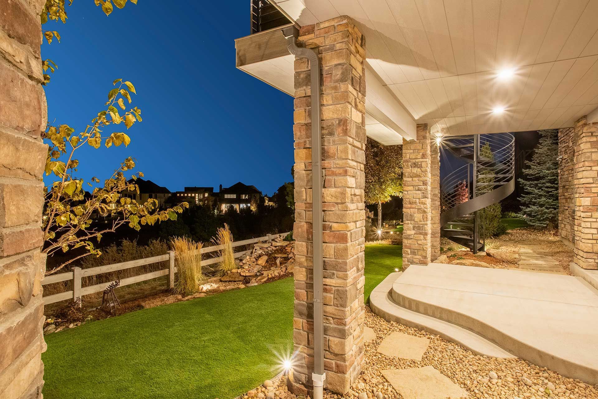 A stone-pillared patio at twilight, overlooking a manicured lawn, a white fence, and distant suburban houses.