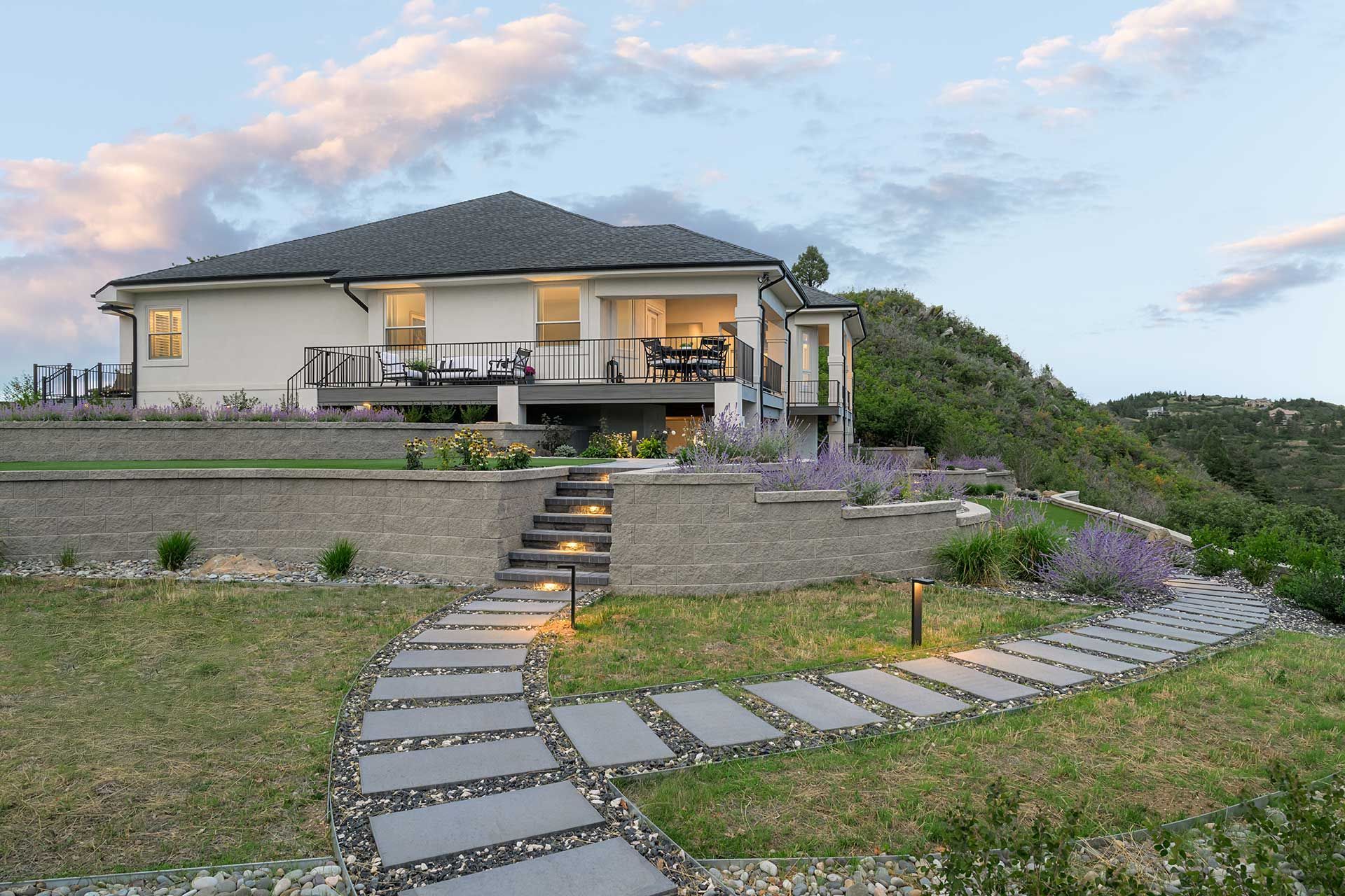 A modern white house on a terraced hillside with a stone walkway leading to the patio under a sunset sky.
