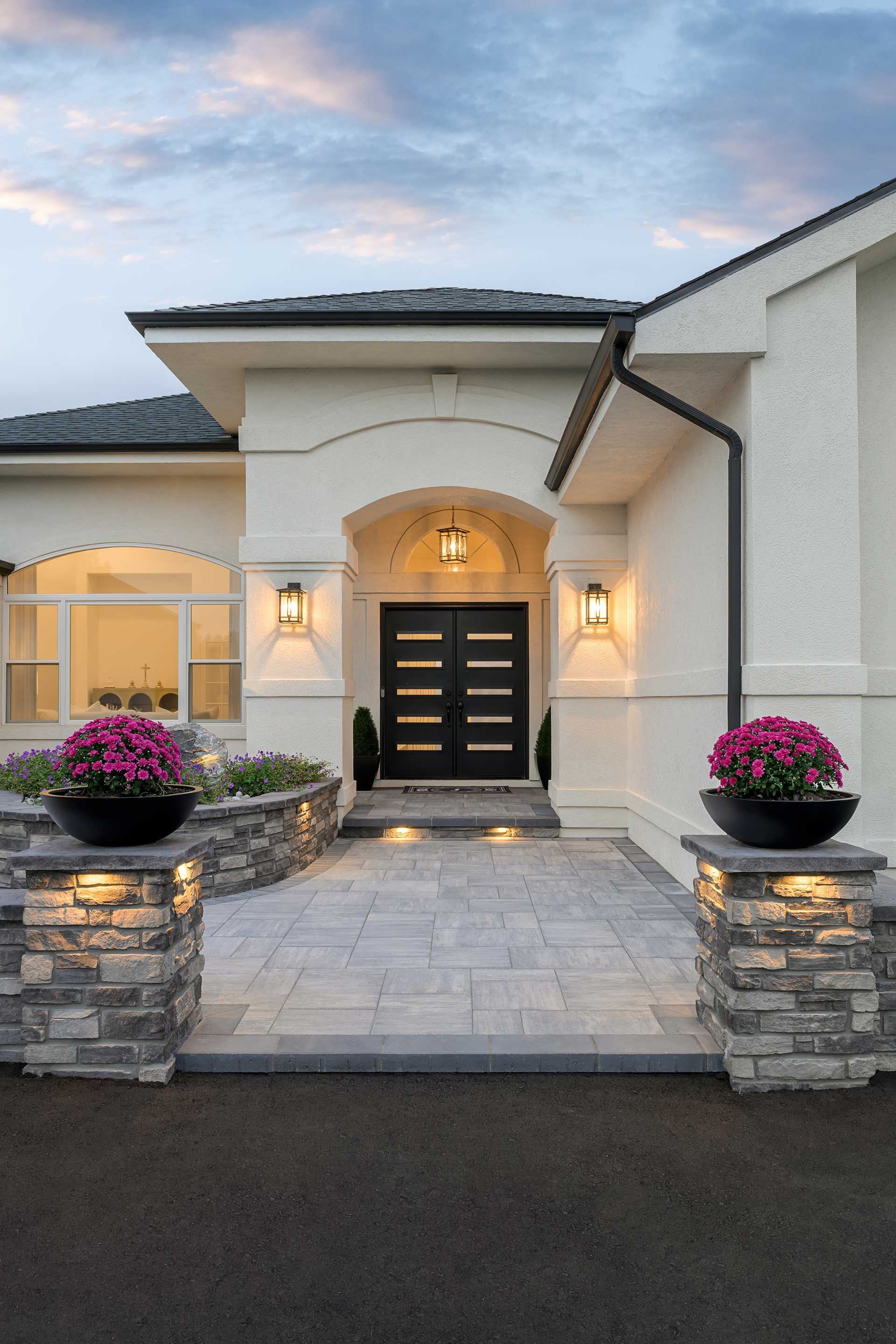 A modern cream-colored house entrance with a stone-paved walkway, dark double doors, and potted purple flowers on pillars.
