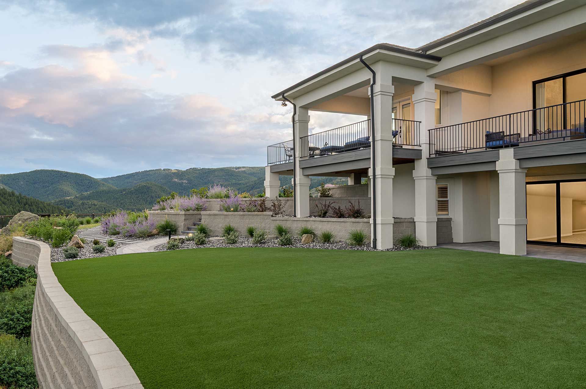 A modern two-story home with a balcony overlooks a manicured green lawn, tiered stone walls, and rolling hills at sunset.
