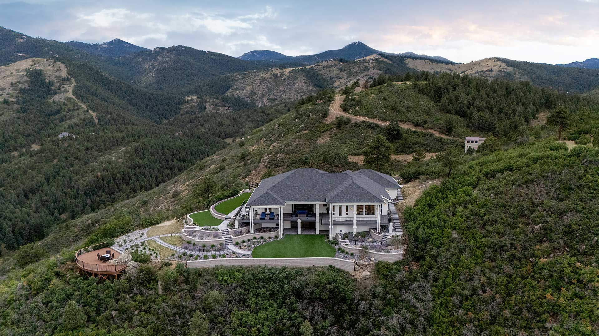 An aerial view of a large white mansion with a dark roof, surrounded by mountains and greenery with an outdoor patio.