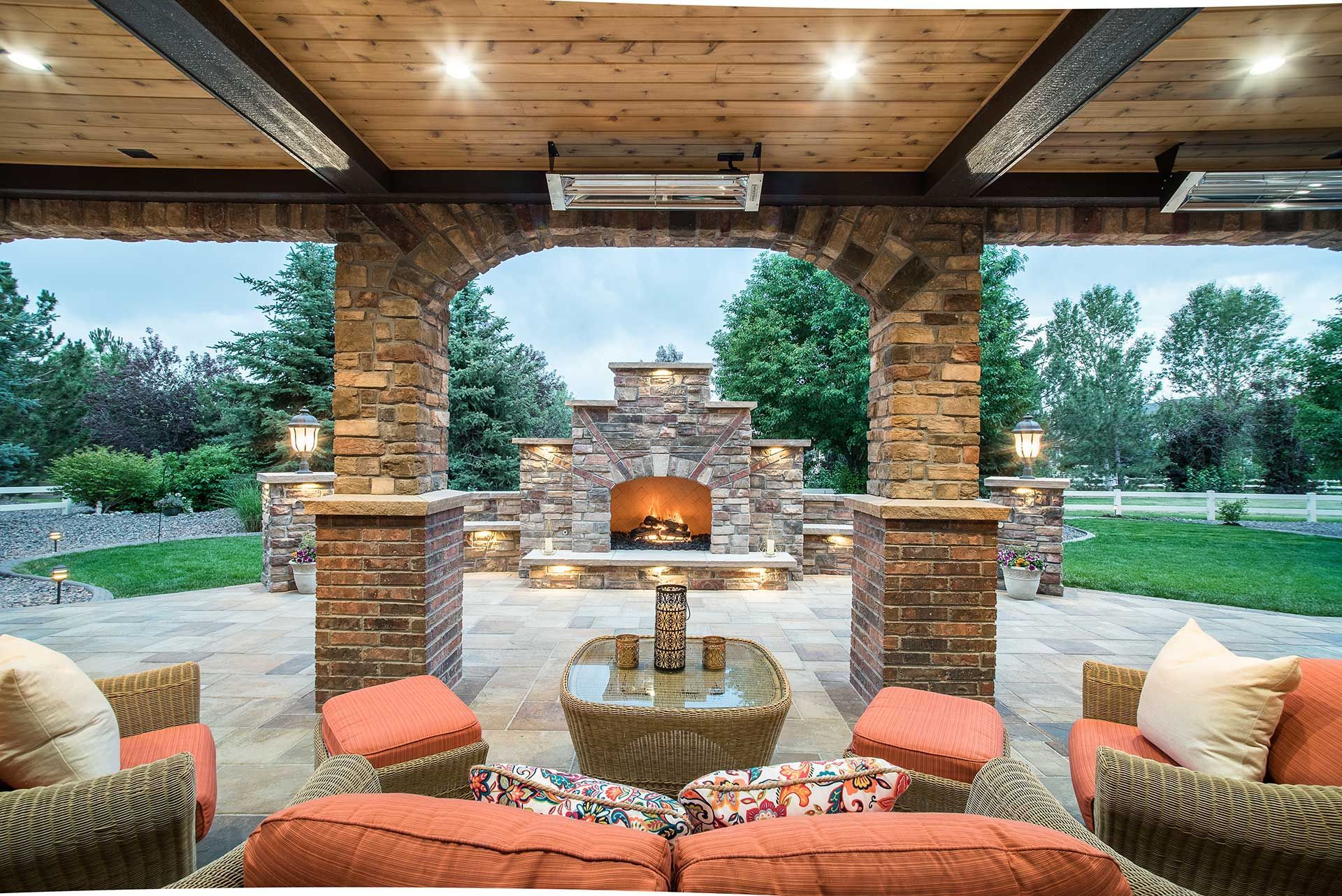 Outdoor patio featuring a stone fireplace under a wooden roof, with wicker furniture and orange cushions, facing a yard.