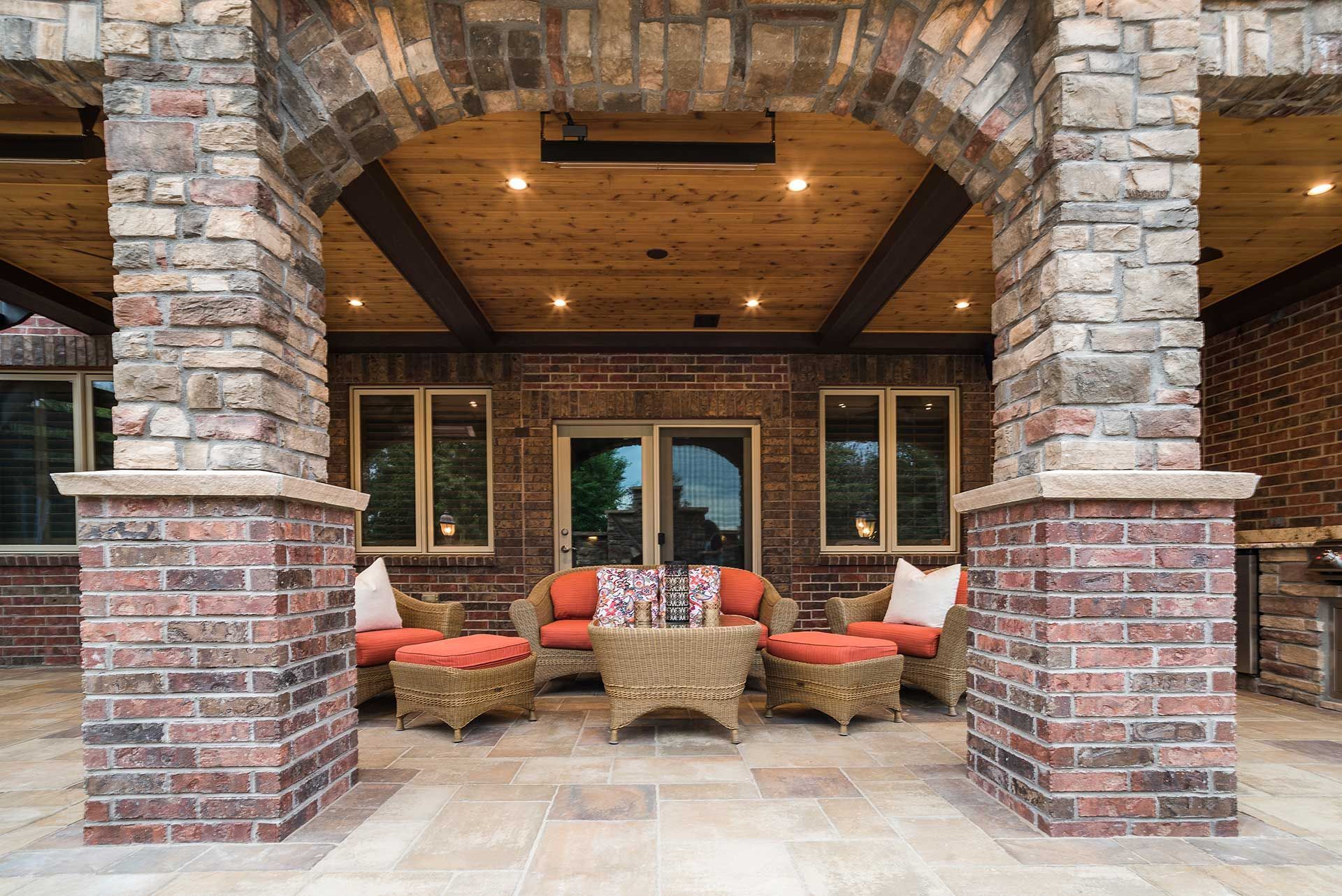 A covered stone patio with rustic brick pillars, wicker furniture, orange cushions, and wood-paneled ceilings.