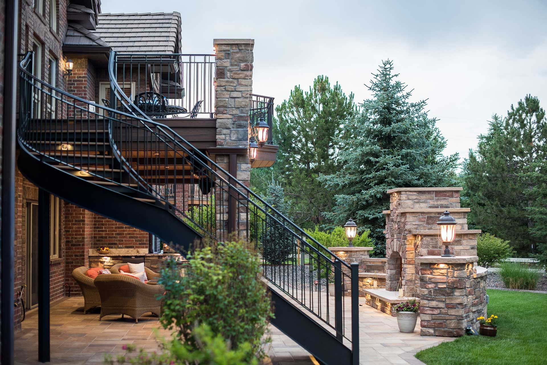 A stone-clad house exterior with a curved metal staircase, patio furniture, and an outdoor stone fireplace in a garden.