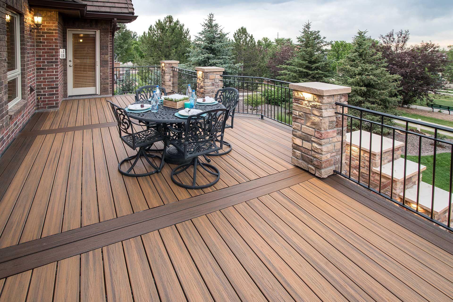 A wooden deck featuring a round metal dining table with chairs, stone-pillar railing, and a brick house exterior.