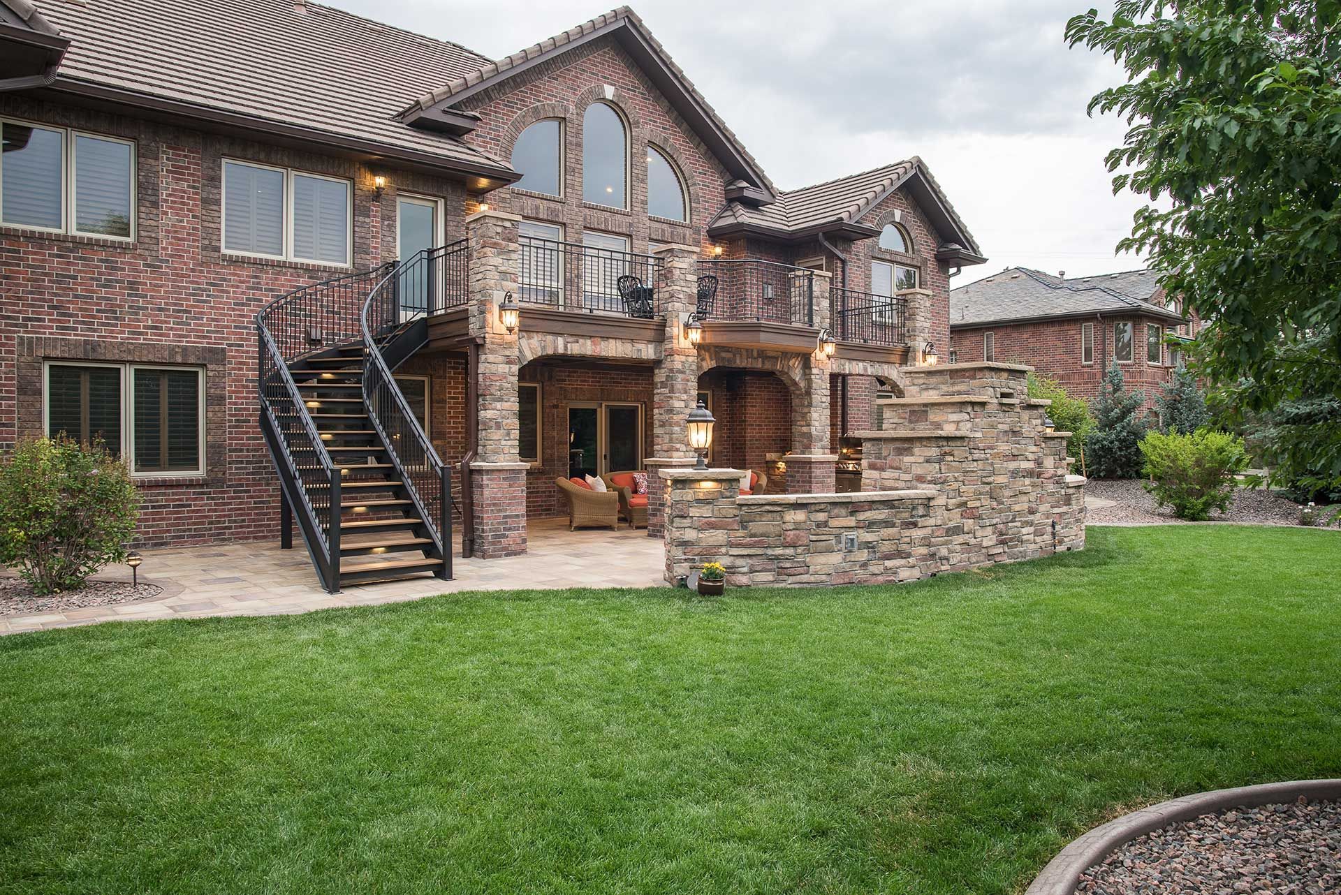 A multi-story brick house with a stone patio, elevated deck, black metal staircase, and a lush green backyard.