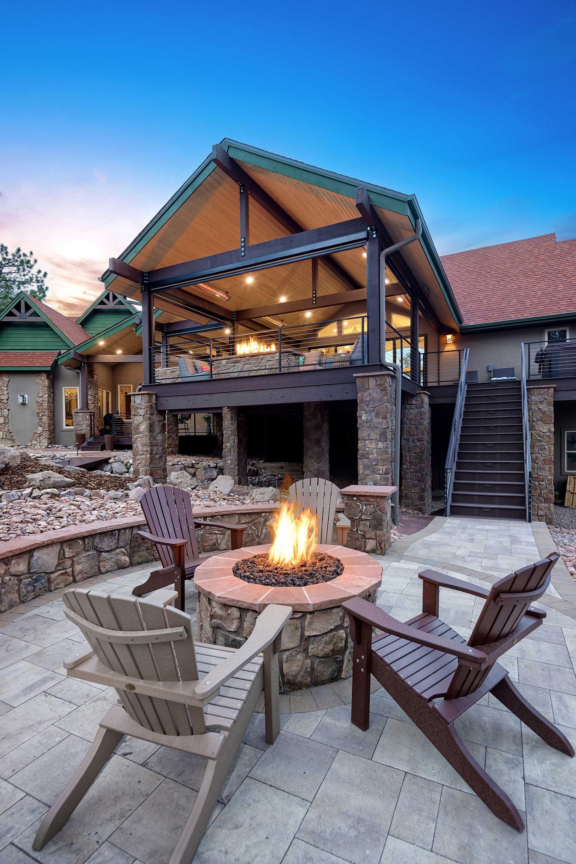 A stone fire pit on a paver patio with three chairs, in front of a rustic house with an elevated covered porch at sunset.