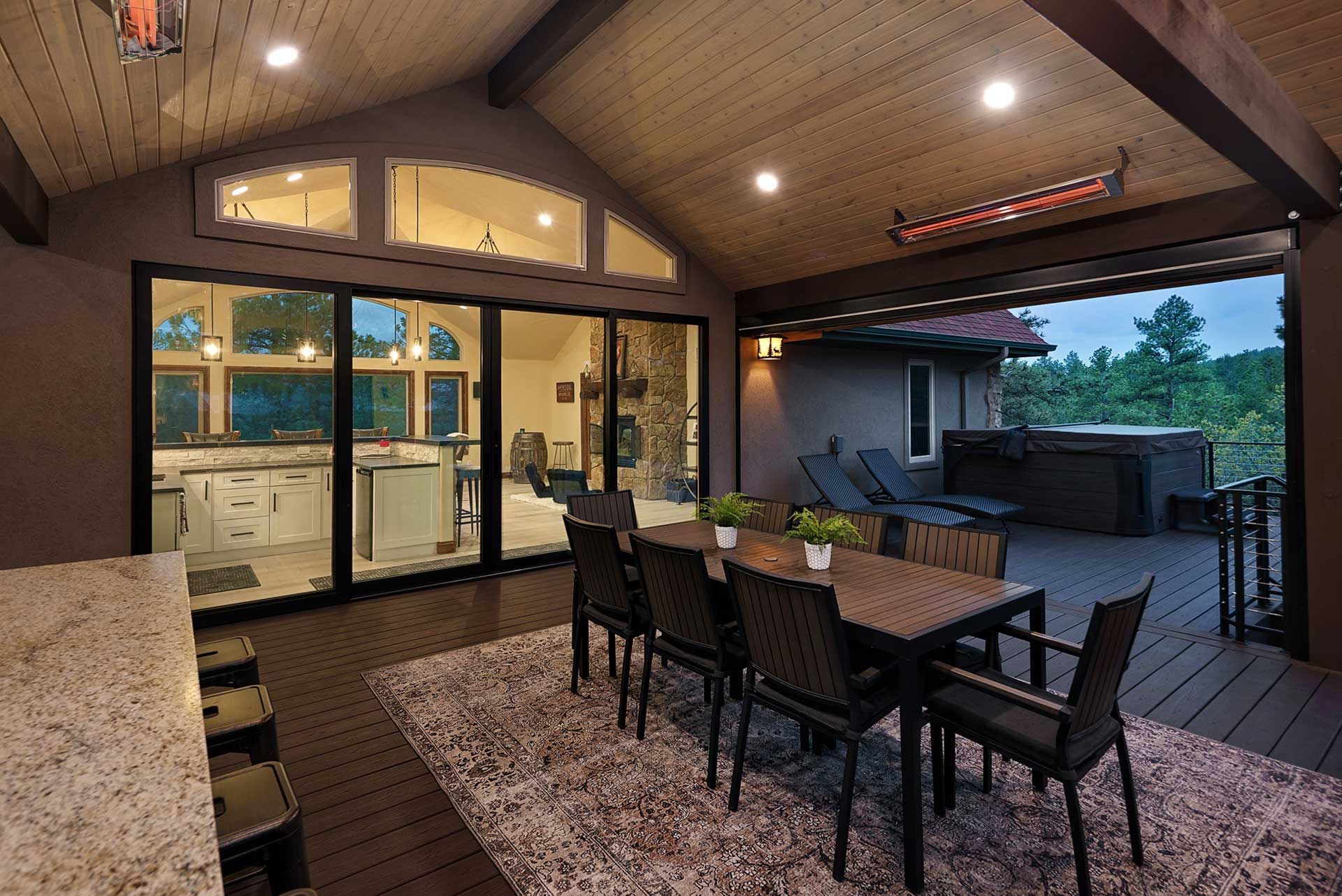 A covered deck features a dining set on a rug, a granite bar, glass sliding doors to an interior, and a hot tub.