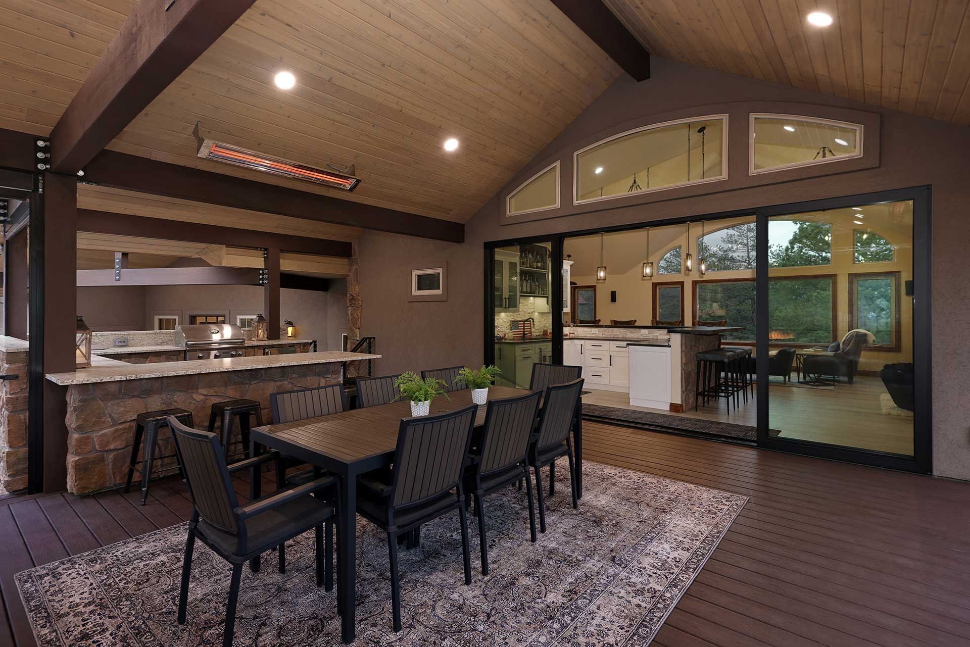 Outdoor dining set with dark chairs on a patterned rug under a covered wooden patio next to a glass-walled indoor room.