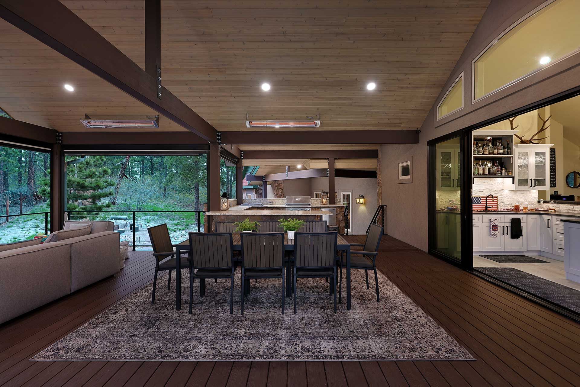 A modern covered deck featuring a dining area with six chairs, an outdoor sofa, and an open view into a kitchen.