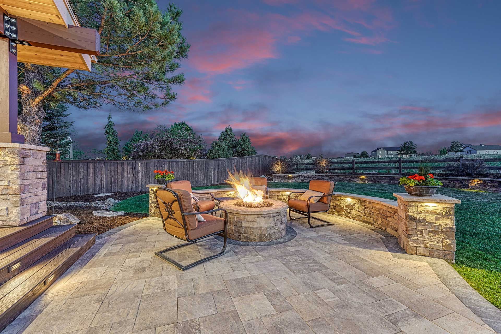 An outdoor stone patio at twilight with a lit fire pit, arranged chairs, and a retaining wall under a purple sky.