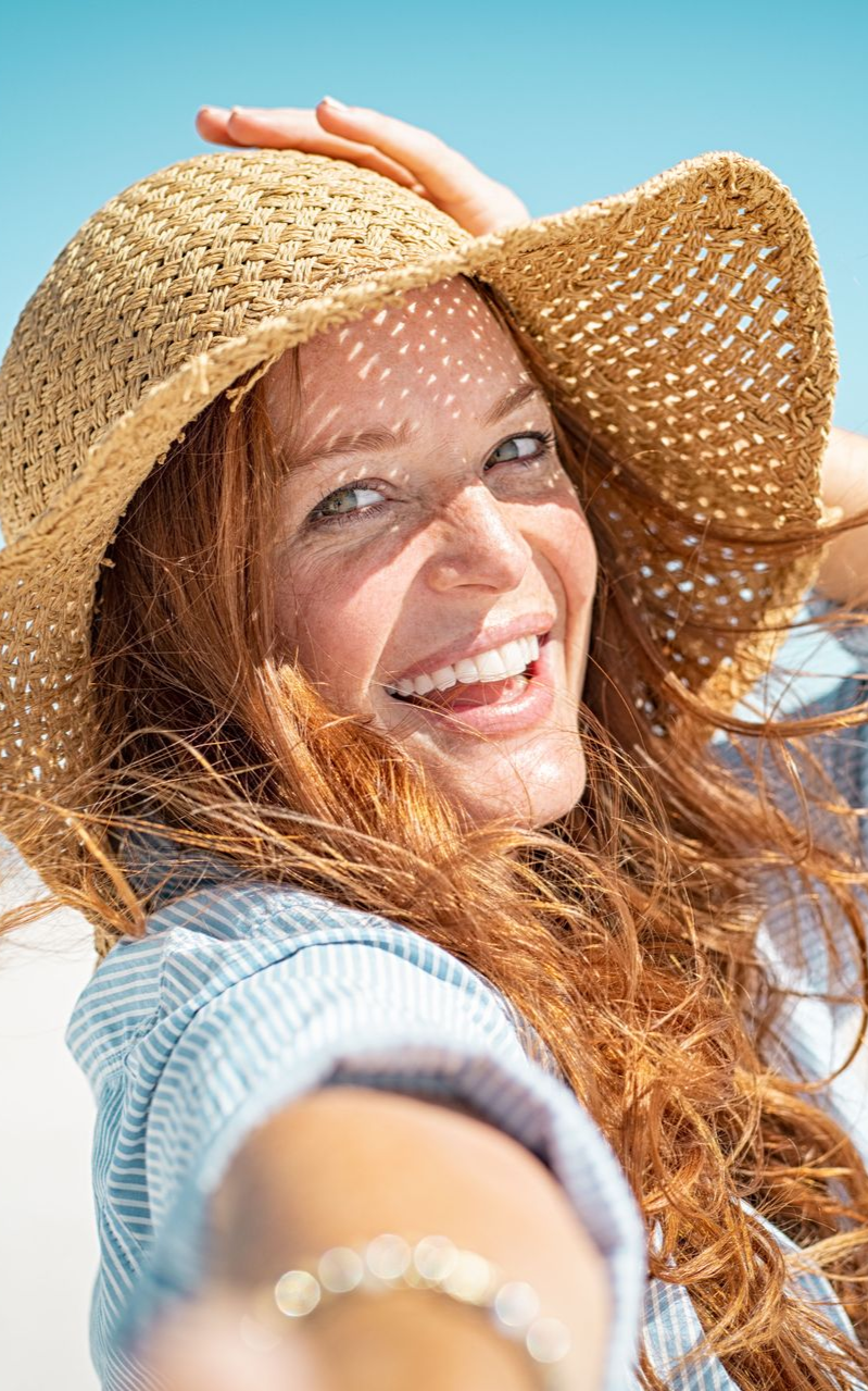 Woman with red hair wearing a straw hat, smiling on a sunny beach.