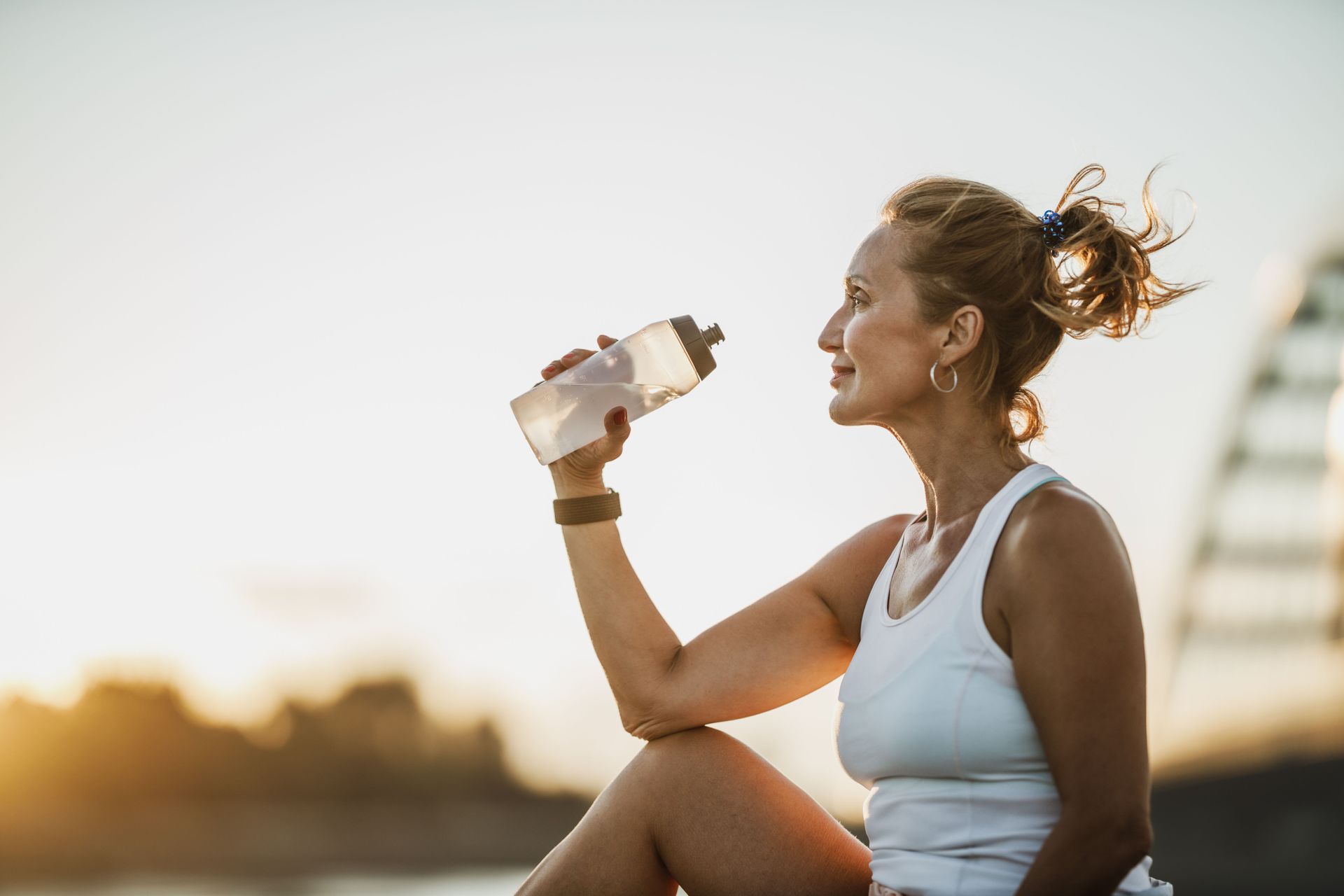 Woman in athletic wear drinks from water bottle, outdoors at sunset.