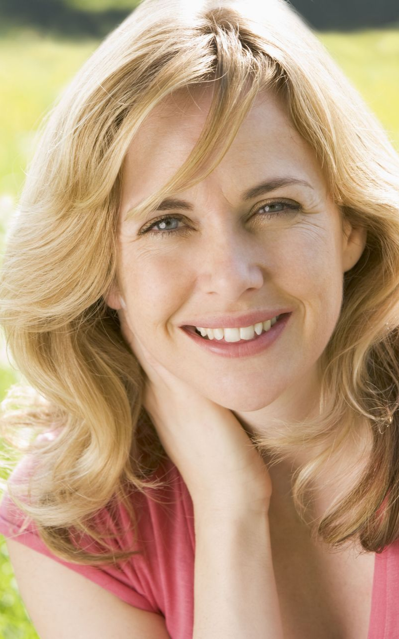 Blonde woman smiling, hand on neck. Wearing pink shirt. Outdoors, blurred green background.