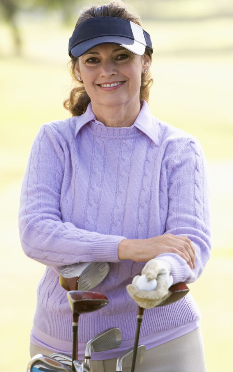 Woman wearing visor, holding golf clubs, smiling on a golf course. She's wearing a purple sweater and khaki pants.