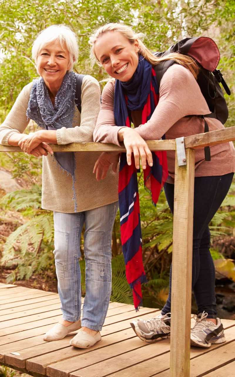 Two women smiling on a wooden bridge in a forest setting. One wears a backpack and scarf.