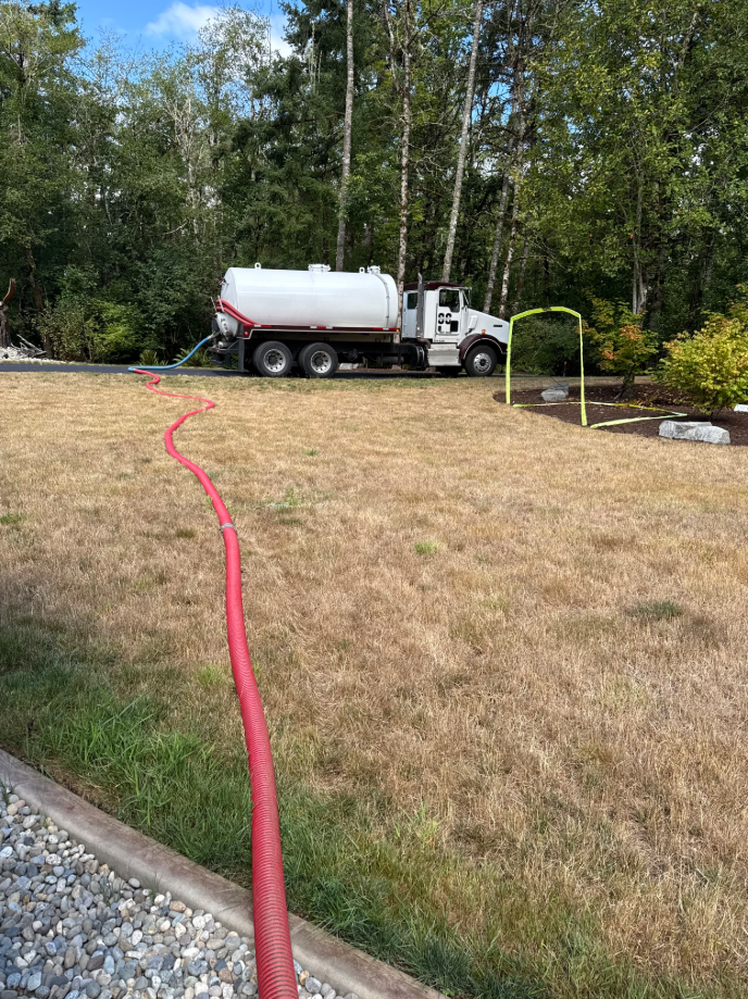 Septic truck with red hose on grassy area. Trees and shrubs in background.