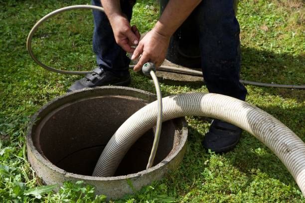 A person holding a hose over an open septic tank in a grassy area, preparing to pump it.