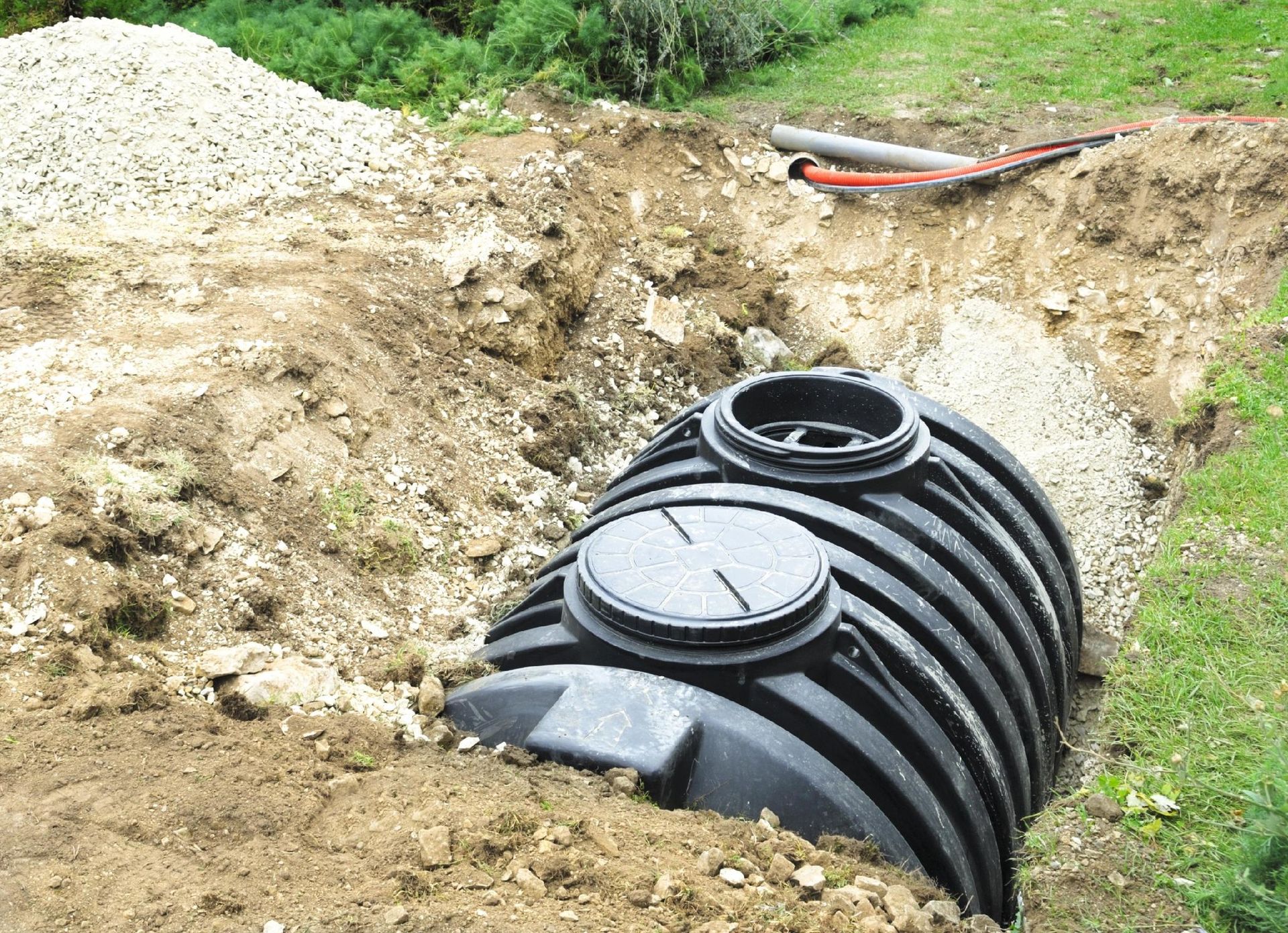 Black septic tank in a trench, partially buried with gravel and dirt, near a grassy area.