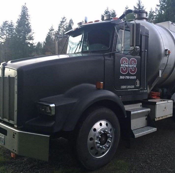 Black tanker truck with silver tank parked outdoors. The truck has a visor and company logo on the door.