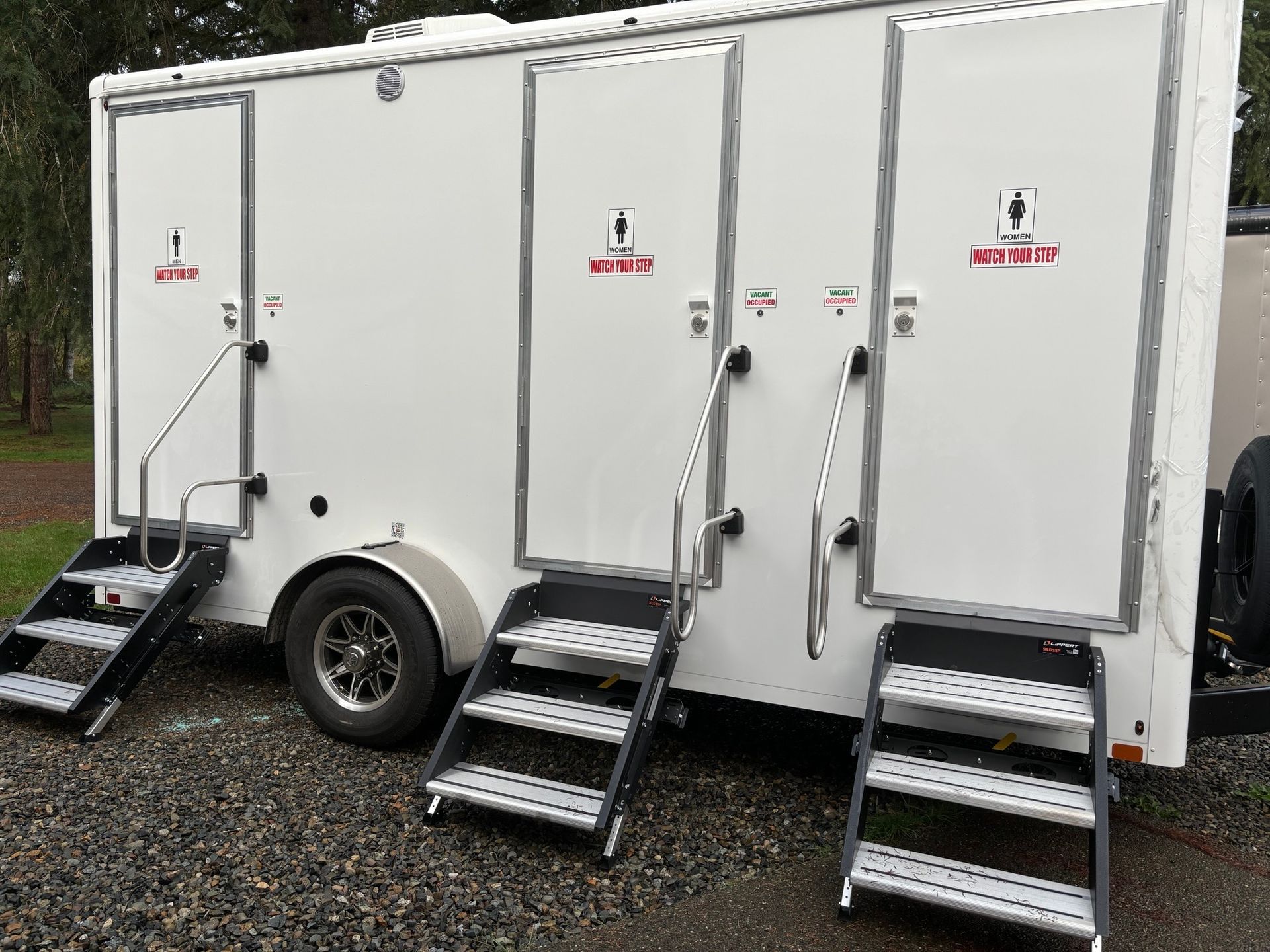 Three-stall portable restroom trailer; white with silver trim and steps, parked outdoors.