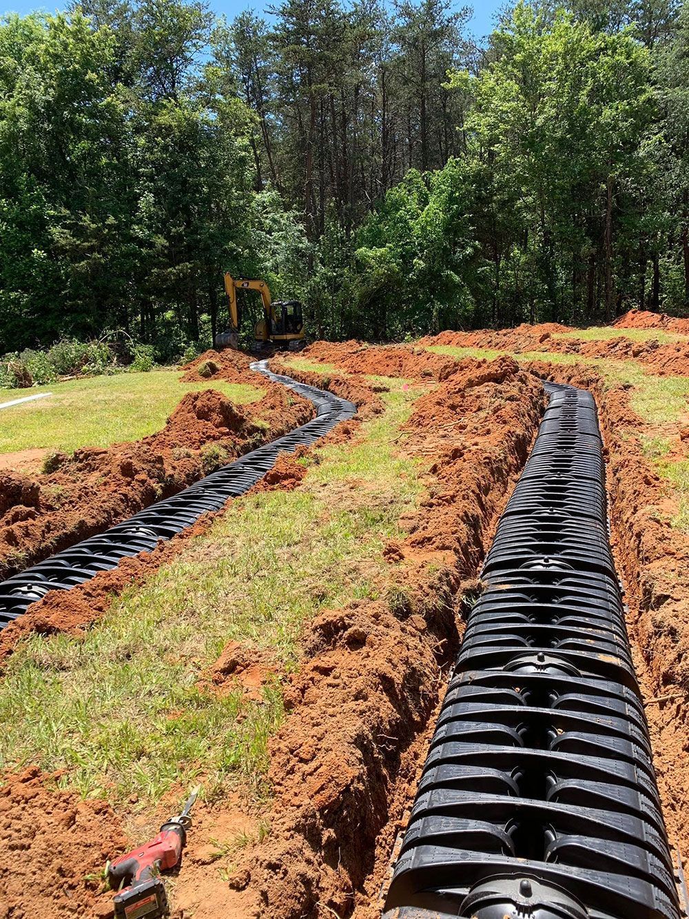 Trenches with black drainage pipes being installed in a grassy area, excavator in the background.