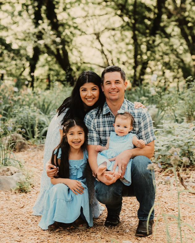 Family of four posing outdoors in a wooded area, with two children seated in front and smiling parents behind.