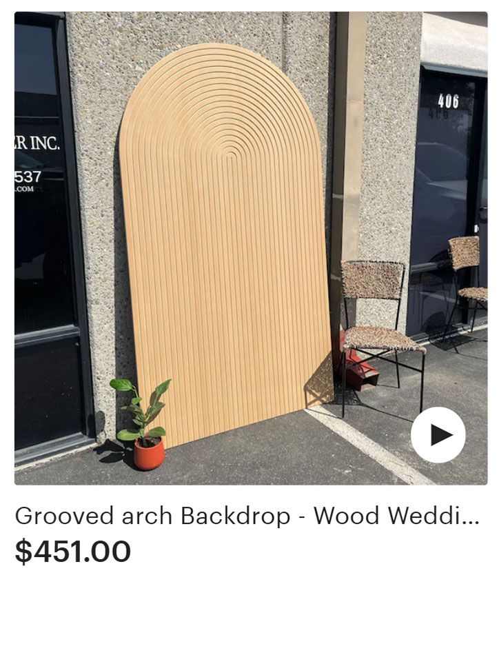 Grooved wood arch backdrop with vertical lines, leaned against a building, next to a plant and chairs.