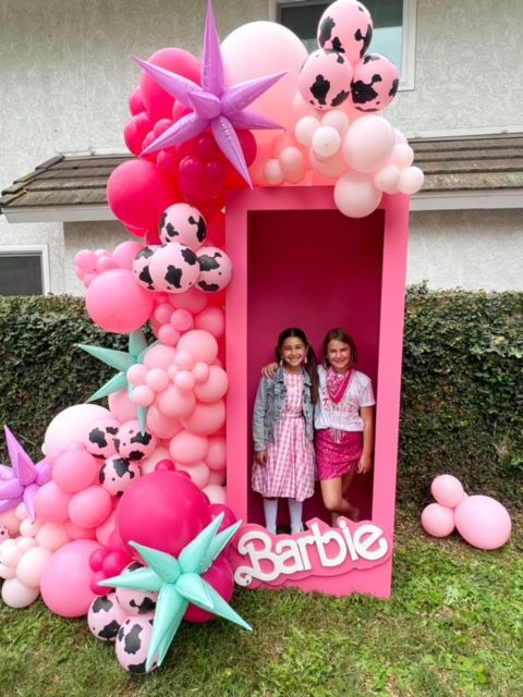 Two girls are standing in a pink barbie box surrounded by pink balloons
