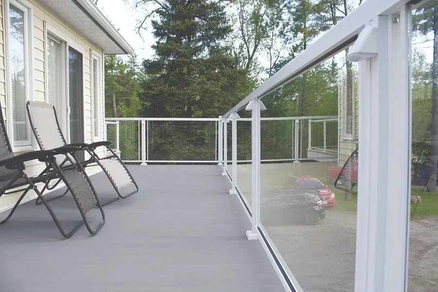 A deck with gray flooring, glass railings, and two lounge chairs.  A car is reflected in the glass.