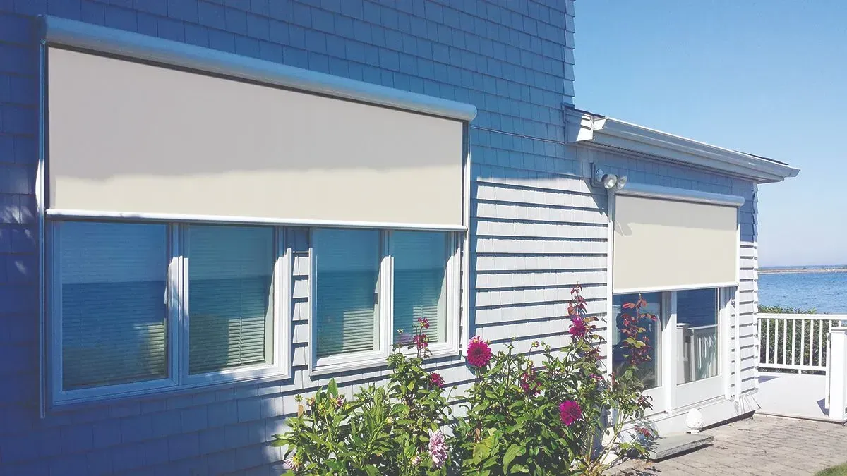 Two outdoor beige roller shades covering windows on a gray house near water.