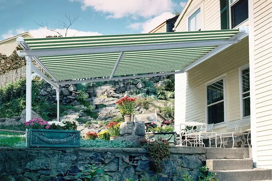 Patio with green and white striped awning, white supports, next to a beige house, stone wall and garden.